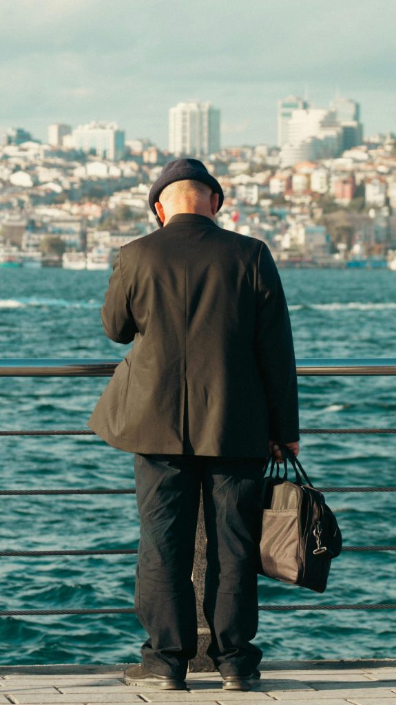 A man in a black suit stands on a pier, gazing at the sea and cityscape ahead.
