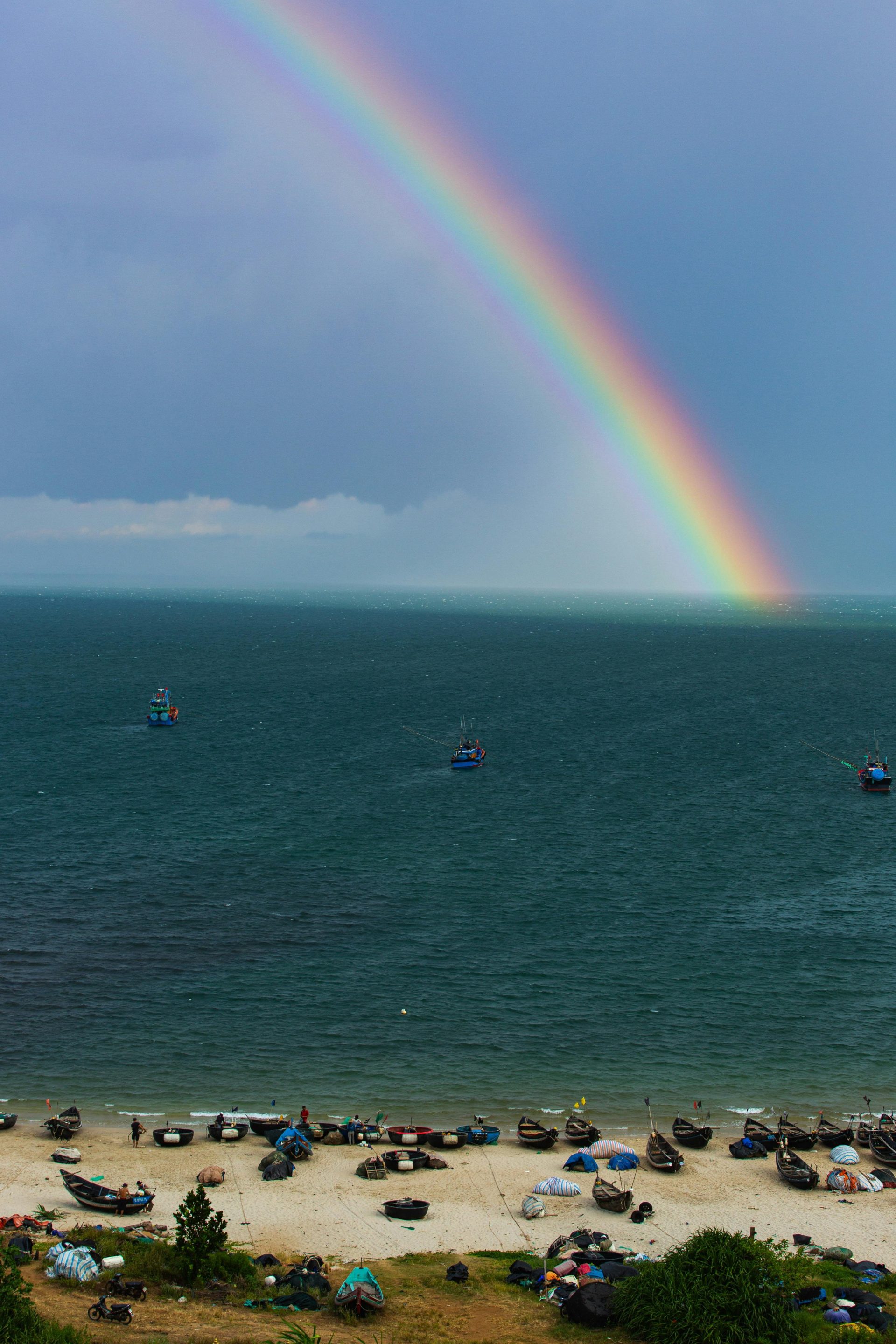 Stunning rainbow arches over a serene beach with fishing boats, capturing nature's beauty.
