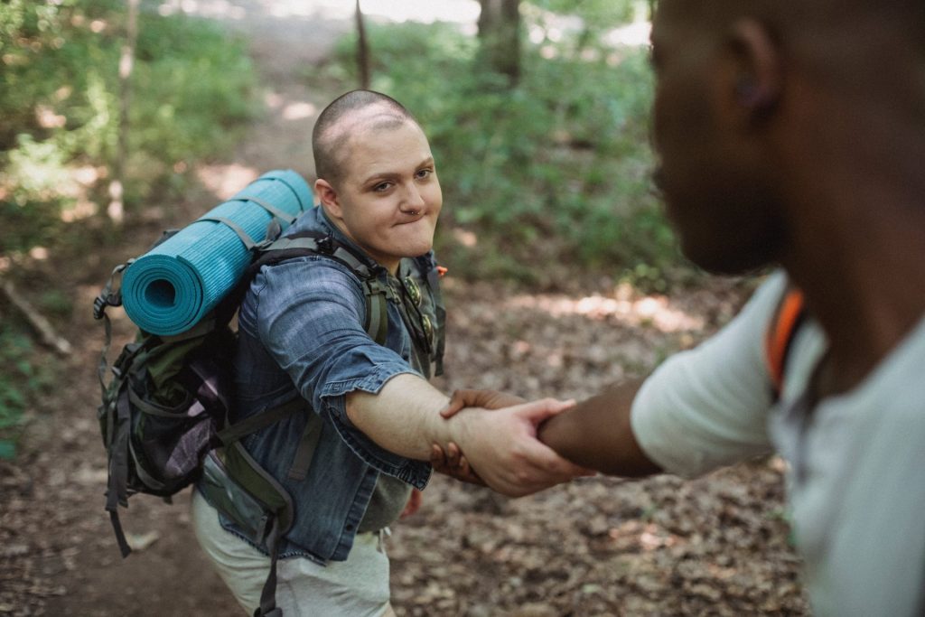 Crop anonymous African American traveler helping friend with backpack with hiking equipment to climb up in forest